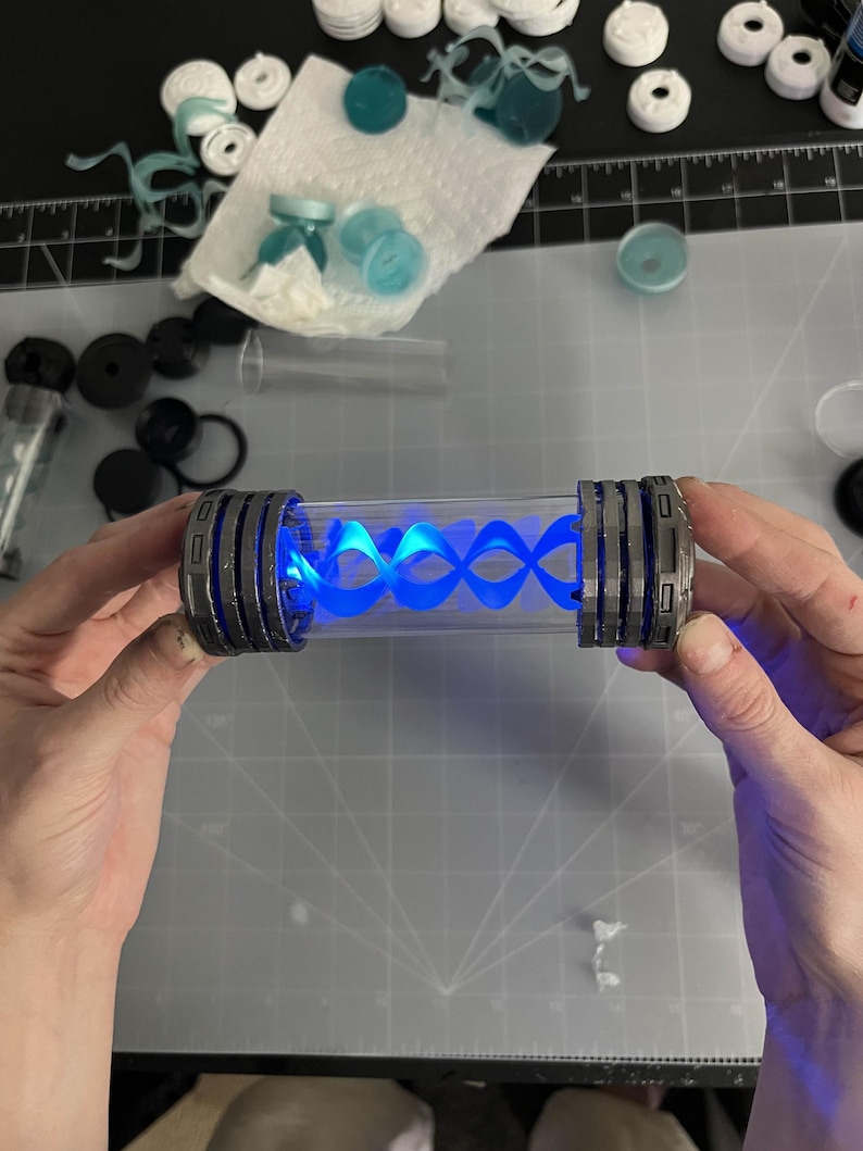 May include: A clear, cylindrical container with gray and silver accents on each end. The container holds a glowing blue, spiral-shaped light. The item is held by two hands against a gray cutting mat.