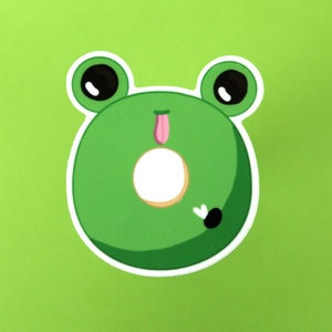 May include: A green frog-shaped donut sticker with white outlines, black eyes, and a pink tongue sticking out.