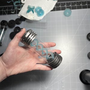 May include: A clear glass vial with a silver metal cap and base, containing a light blue, swirling, spiral-shaped design. The vial is held in a hand, with various other small, light blue and white components scattered around on a gray surface.