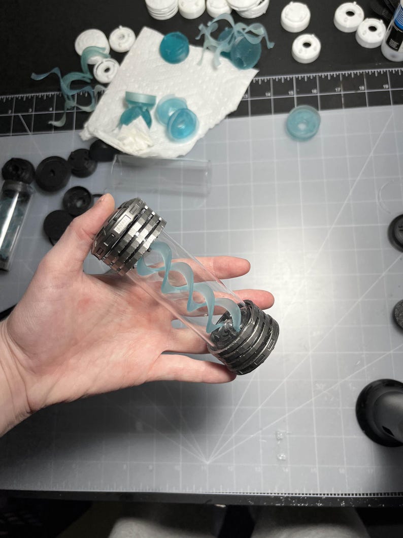 May include: A clear glass vial with a silver metal cap and base, containing a light blue, swirling, spiral-shaped design. The vial is held in a hand, with various other small, light blue and white components scattered around on a gray surface.