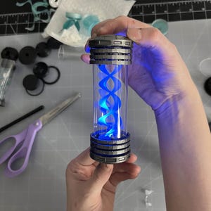 May include: A cylindrical, clear container with a blue, double helix design illuminated by blue light. The container has a metallic gray top and bottom with a ribbed design. The item is held in a person's hands.