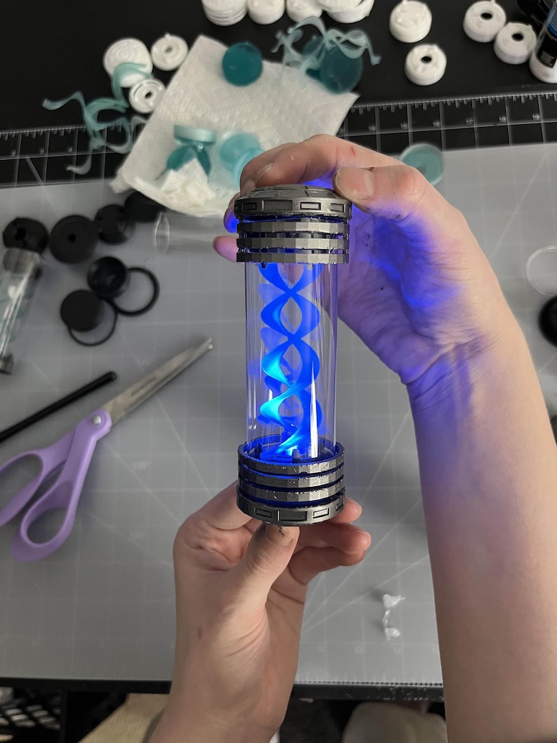 May include: A cylindrical, clear container with a blue, double helix design illuminated by blue light. The container has a metallic gray top and bottom with a ribbed design. The item is held in a person's hands.