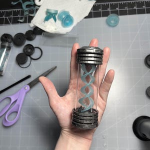 May include: A clear, cylindrical container with a silver-colored, layered cap and base. Inside, a light blue, double helix structure is visible. The container is held in a hand, with various crafting supplies in the background.