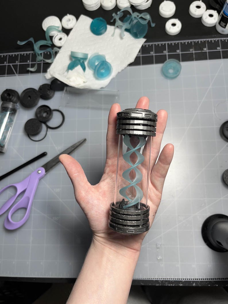 May include: A clear, cylindrical container with a silver-colored, layered cap and base. Inside, a light blue, double helix structure is visible. The container is held in a hand, with various crafting supplies in the background.