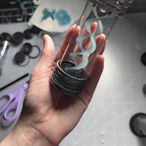May include: A clear glass vial with a silver cap and base, containing a light blue, swirling, double helix design. The vial is held in a hand, with other crafting supplies visible in the background.