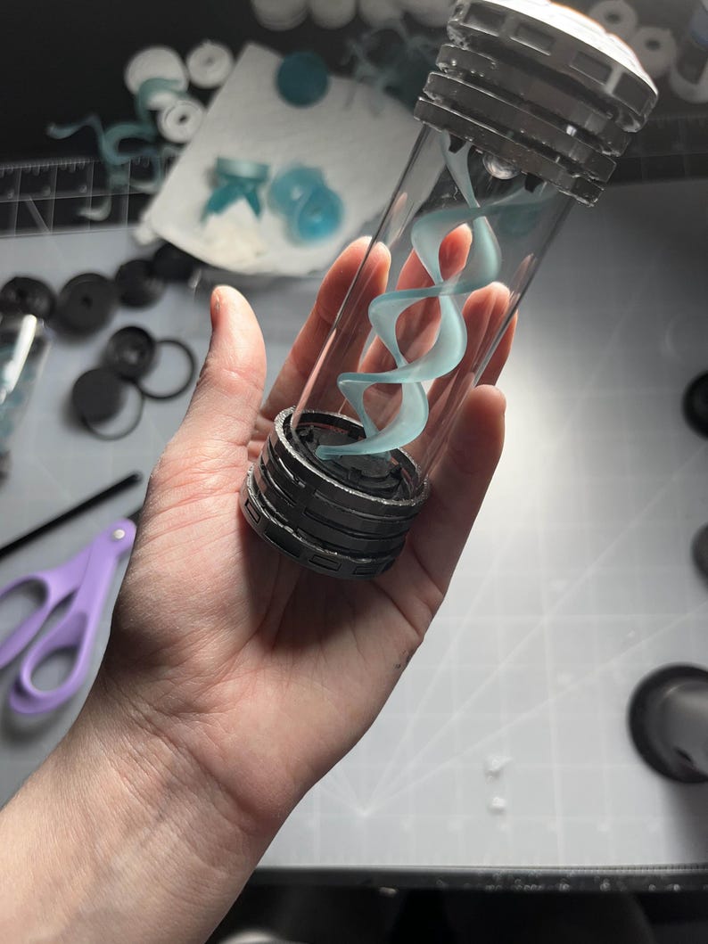 May include: A clear glass vial with a silver cap and base, containing a light blue, swirling, double helix design. The vial is held in a hand, with other crafting supplies visible in the background.