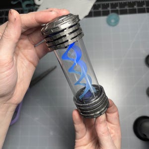 May include: A clear, cylindrical vial with a metallic, ribbed cap and base, containing a glowing blue, double helix design. The vial is held in two hands, with other crafting supplies visible in the background.