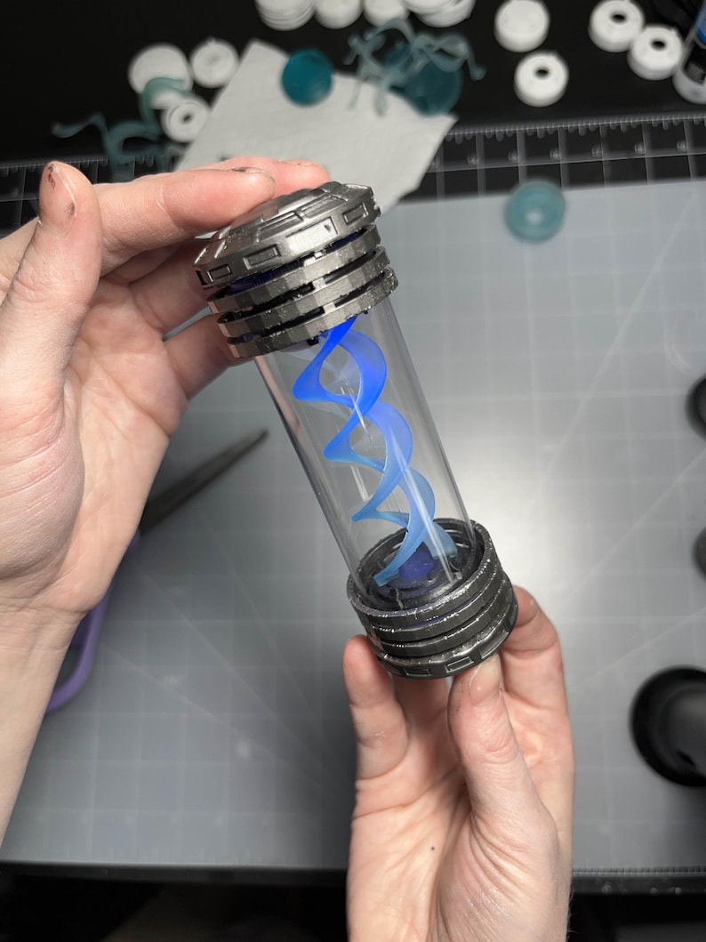 May include: A clear, cylindrical vial with a metallic, ribbed cap and base, containing a glowing blue, double helix design. The vial is held in two hands, with other crafting supplies visible in the background.