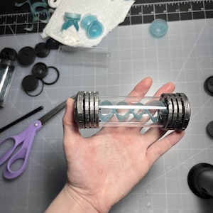 May include: A clear, cylindrical container with a silver-colored, ridged cap on each end. Inside, a light blue, spiral-shaped object is visible. Various other small, light blue and white components are scattered around the container.