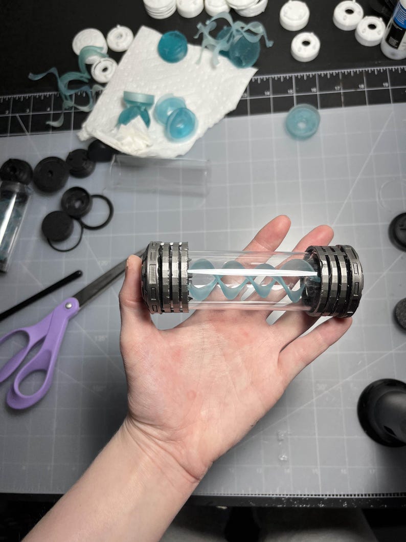 May include: A clear, cylindrical container with a silver-colored, ridged cap on each end. Inside, a light blue, spiral-shaped object is visible. Various other small, light blue and white components are scattered around the container.