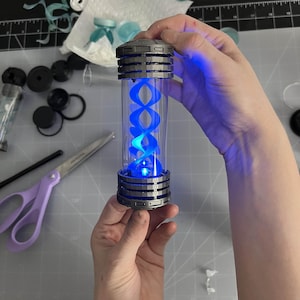 May include: A cylindrical, clear container with a blue, glowing, double helix design inside. The container has a metallic gray top and bottom with a ribbed design. The object is held in a person's hands, with other crafting supplies in the background.