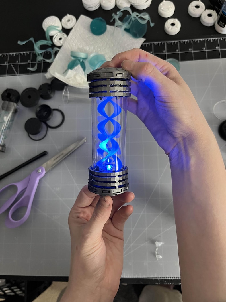 May include: A cylindrical, clear container with a blue, glowing, double helix design inside. The container has a metallic gray top and bottom with a ribbed design. The object is held in a person's hands, with other crafting supplies in the background.