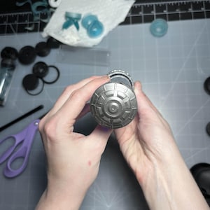 May include: A detailed view of a cylindrical object with a patterned, metallic end cap. The object is held by two hands, with various crafting supplies and tools visible in the background, including scissors and small plastic components.
