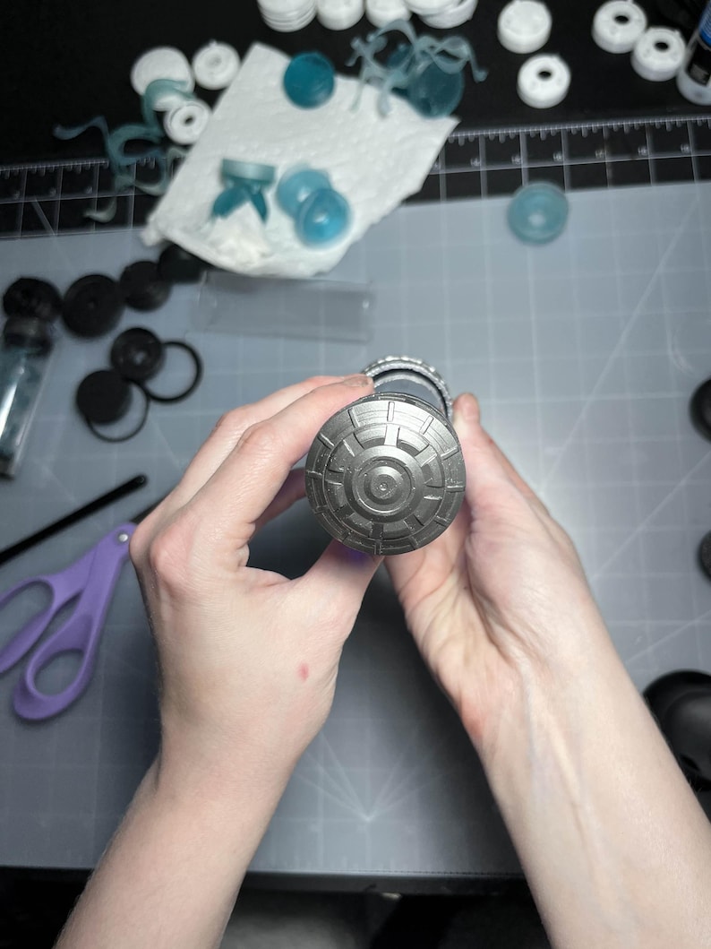 May include: A detailed view of a cylindrical object with a patterned, metallic end cap. The object is held by two hands, with various crafting supplies and tools visible in the background, including scissors and small plastic components.