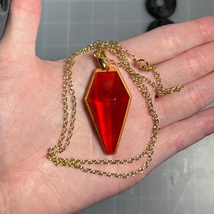 May include: A gold-coloured chain necklace with a pendant. The pendant is a red, coffin-shaped gem with a gold-coloured border. The necklace is displayed on a person's open palm.