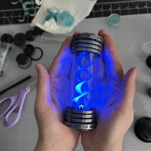 May include: A cylindrical glass vial with a silver and black metallic cap and base, illuminated by a bright blue light. The vial contains a blue, double helix structure. The image is of a science fiction or fantasy prop.