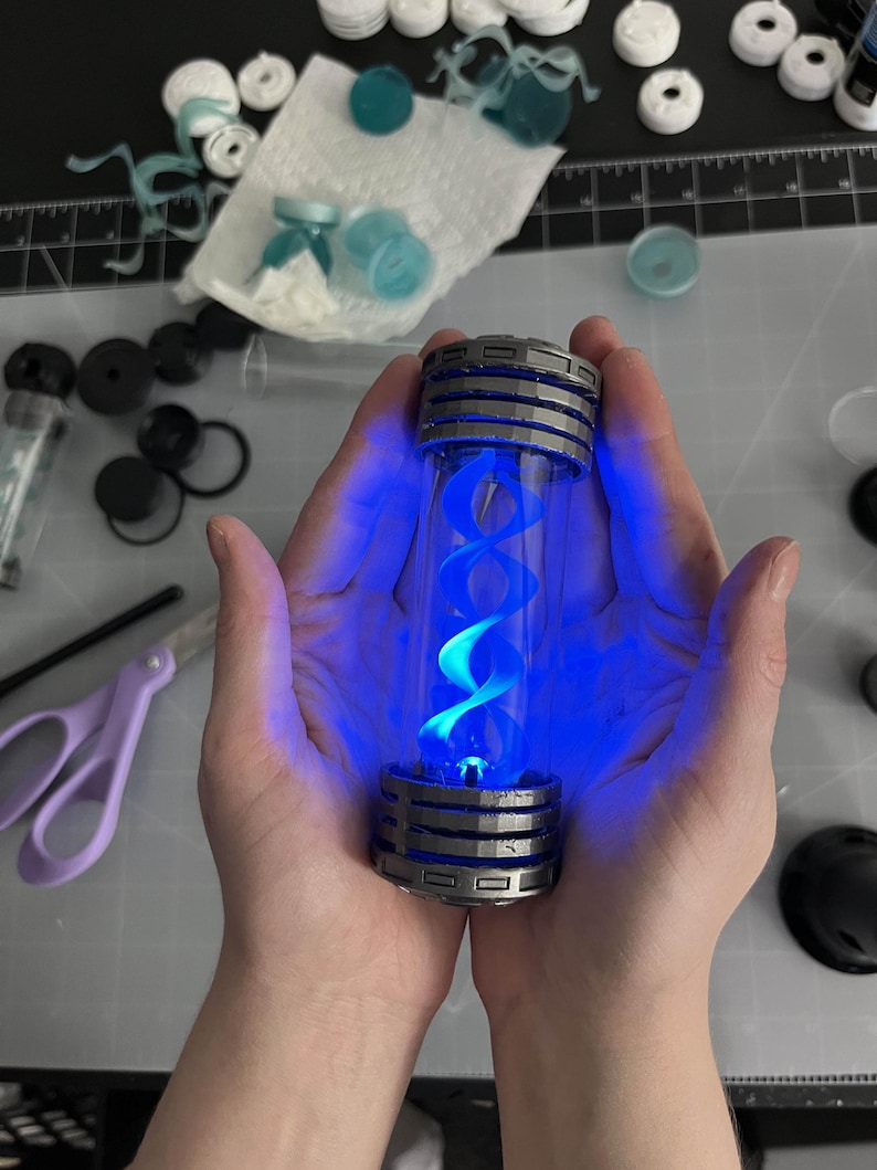 May include: A cylindrical glass vial with a silver and black metallic cap and base, illuminated by a bright blue light. The vial contains a blue, double helix structure. The image is of a science fiction or fantasy prop.