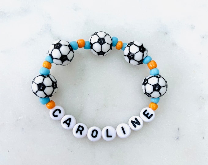 May include: A beaded bracelet featuring five spherical soccer ball beads interspersed with small light blue and orange round beads. The bottom section of the bracelet consists of white circular beads with the black printed name CAROLINE.