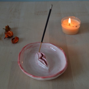 May include: A pink ceramic incense holder with a stylized design, holding a lit incense stick. A small candle burns nearby, with dried flowers scattered around. The holder has a pink rim and sits on a light wooden surface.