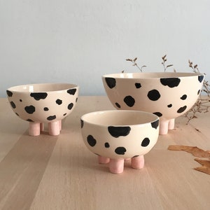 May include: Three ceramic bowls with black and white spotted patterns and pink legs. The bowls are nested inside each other, with the largest bowl on the bottom and the smallest bowl on top.