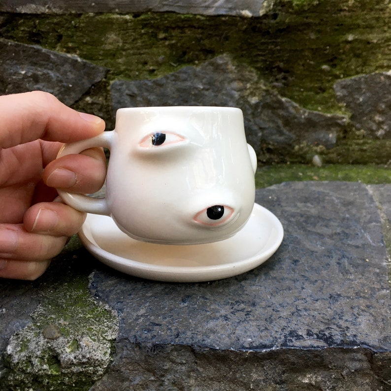 May include: White ceramic coffee cup with two large eyes and a saucer. The eyes are black with pink eyelids.