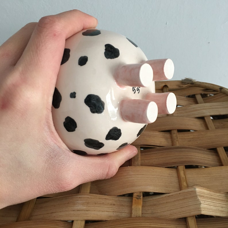 May include: A ceramic piggy bank with a white base and black spots, resembling a cow. The piggy bank has three pink legs with white hooves. The piggy bank is being held in a hand.