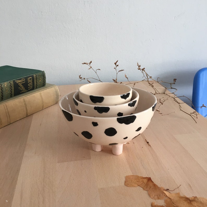 May include: Set of three ceramic bowls with a black and white cow print design. The largest bowl has four small feet.