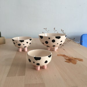 May include: Three ceramic bowls with black and white spotted patterns and pink legs. The bowls are arranged in a size order from smallest to largest.