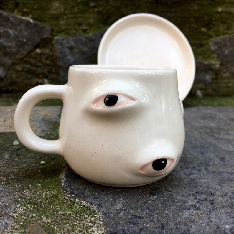 May include: White ceramic mug with two large eyes, one on each side. The eyes have black pupils and pink eyelids. The mug has a rounded shape and a handle.