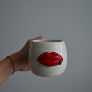 Könnte beinhalten: Weiße Keramiktasse mit einem roten, dreidimensionalen Lippen-Design. Die Lippe ist leicht geöffnet und hat einen schwarzen Strich in der Mitte.