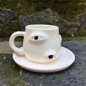 May include: White ceramic coffee cup with two black eyes and pink eyelids. The cup is on a white saucer.