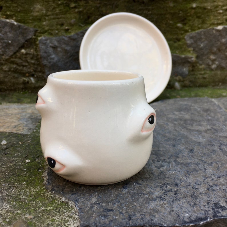 May include: White ceramic mug with three eyes, each with a black pupil. The mug is sitting on a gray stone surface.