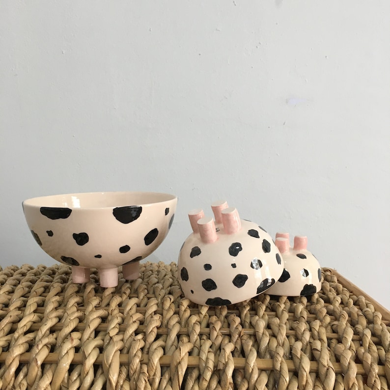 May include: Three ceramic bowls with a pink and black spotted design. The bowls have three legs and are shaped like a cow.