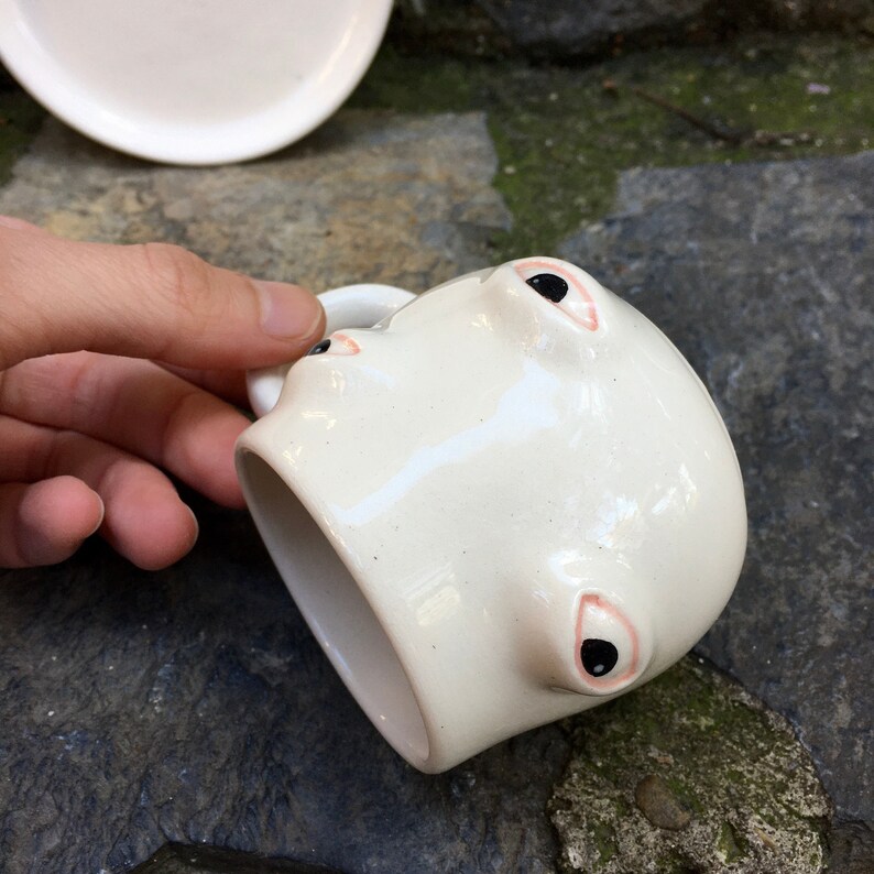 May include: White ceramic mug with two black eyes and pink eyelids. The mug is shaped like a face and has a handle on the side.