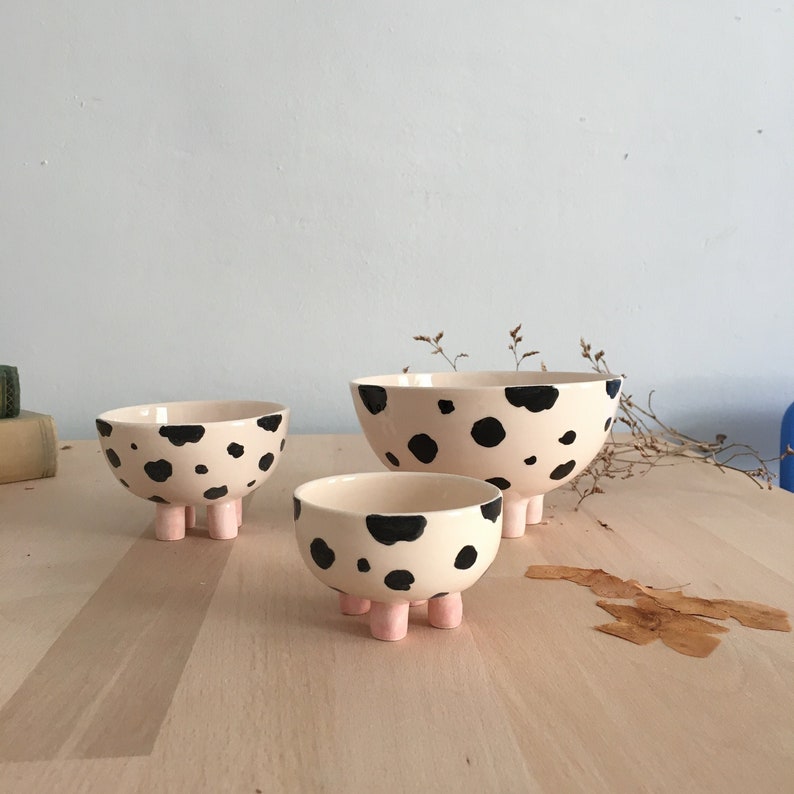 May include: Three ceramic bowls with black and white spotted patterns and pink legs. The bowls are nested inside each other, with the largest bowl on the bottom and the smallest bowl on top.