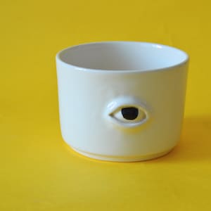 Peut inclure: Une tasse en céramique blanche avec un motif d'œil noir. La tasse est cylindrique avec une surface lisse et un seul œil sculpté sur le côté. Le fond est de couleur jaune uni.
