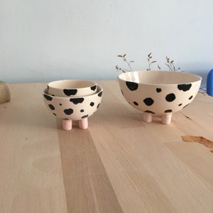 May include: Two ceramic bowls with black and white spots and pink legs. The larger bowl is on the right and the smaller bowl is on the left, stacked on top of the larger bowl.