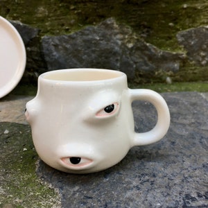 May include: White ceramic coffee mug with three eyes, two on the side and one on the front. The eyes are black with pink eyelids.