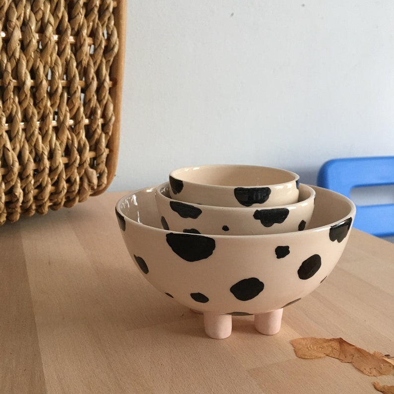 May include: Three ceramic bowls with a black and white cow print design. The largest bowl has three short legs and is nested inside the other two bowls.