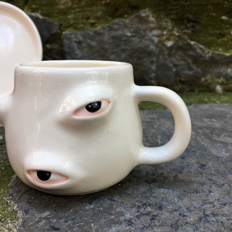 May include: White ceramic coffee mug with two large eyes and a pink eyelid. The mug has a rounded shape and a handle.