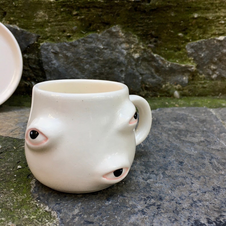 May include: White ceramic coffee mug with three eyes, two on the front and one on the side. The eyes are black with pink eyelids.