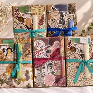 Puede incluir: Cinco paquetes rectangulares envueltos en papel marrón, decorados con varias pegatinas, efímeros de papel y una cinta. Cada paquete incluye un bolígrafo o rotulador y está atado con una cinta de satén de color azul cerceta o magenta.