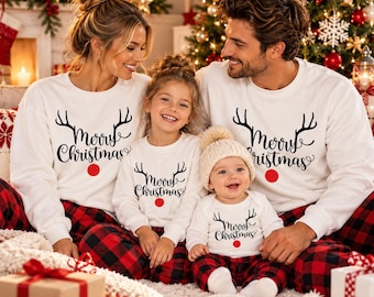 May include: Matching white long-sleeve sweatshirts featuring black reindeer antlers, the text Merry Christmas, and a red nose graphic. The family wears these with red and black buffalo plaid pants. The baby wears a matching white beanie hat.