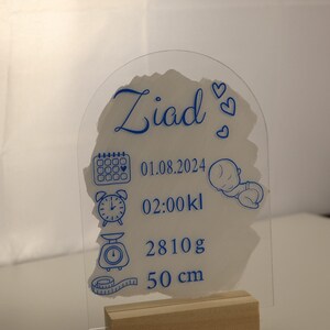 K&ouml;nnte beinhalten: Eine transparente Acryl-Geburtsanzeige mit blauer Beschriftung. Die Plakette zeigt den Namen "Ziad", das Datum "01.08.2024", die Uhrzeit "02:00 kl", das Gewicht "2810g" und die L&auml;nge "50 cm". Die Plakette steht auf einem Holzst&auml;nder.