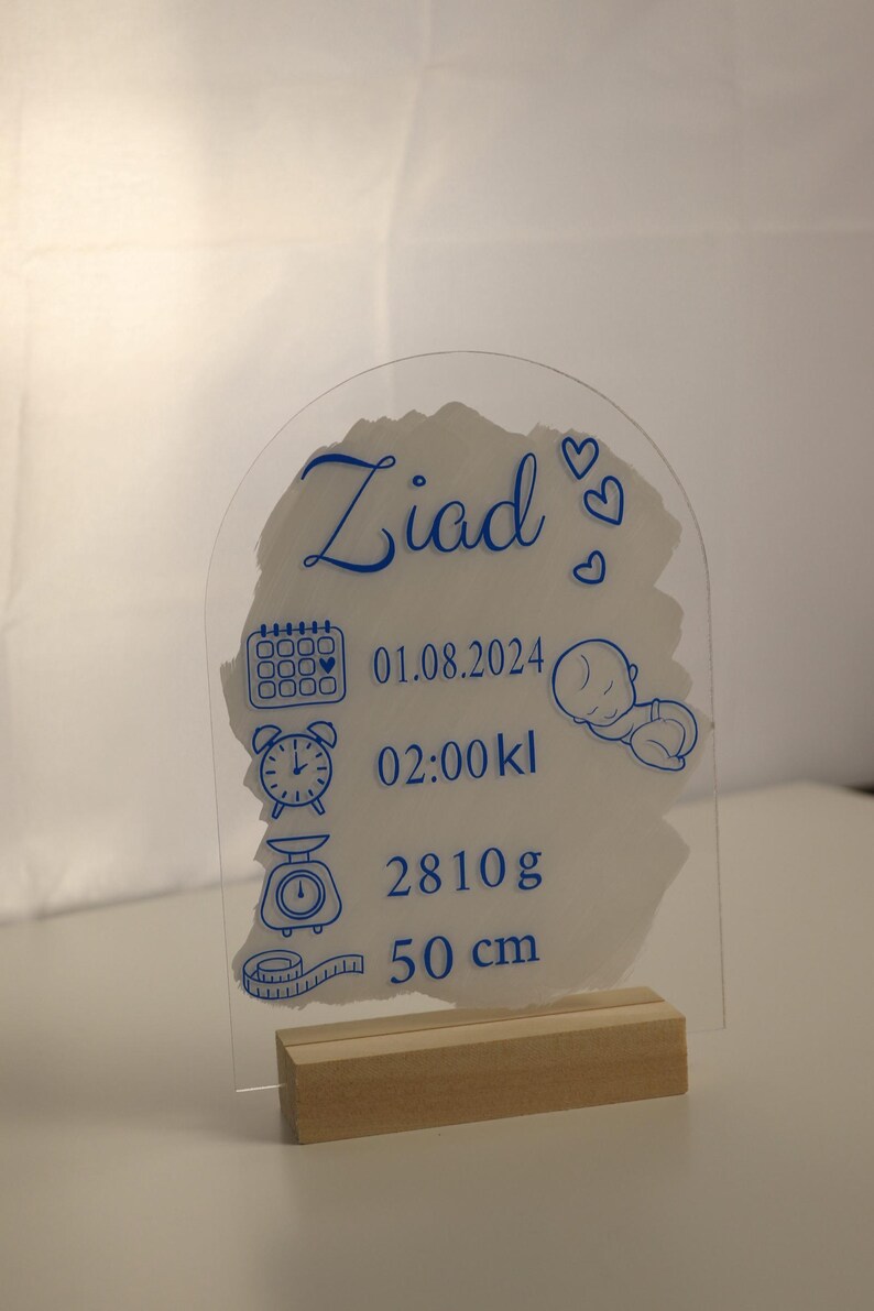 K&ouml;nnte beinhalten: Eine transparente Acryl-Geburtsanzeige mit blauer Beschriftung. Die Plakette zeigt den Namen "Ziad", das Datum "01.08.2024", die Uhrzeit "02:00 kl", das Gewicht "2810g" und die L&auml;nge "50 cm". Die Plakette steht auf einem Holzst&auml;nder.