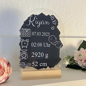 K&ouml;nnte beinhalten: Ein klares Acrylschild mit einem schwarzen Schiefer-Einsatz. Das Schild hat eine wei&szlig;e Aufschrift mit dem Text "Kyan" und enth&auml;lt Symbole f&uuml;r Kalender, Uhr, Waage und Ma&szlig;band. Der Schiefer-Einsatz zeigt das Datum "07.03.2025", die Uhrzeit "02:08 Uhr", das Gewicht "2920 g" und die L&auml;nge "52 cm".