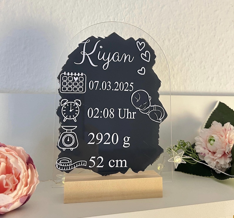 K&ouml;nnte beinhalten: Ein klares Acrylschild mit einem schwarzen Schiefer-Einsatz. Das Schild hat eine wei&szlig;e Aufschrift mit dem Text "Kyan" und enth&auml;lt Symbole f&uuml;r Kalender, Uhr, Waage und Ma&szlig;band. Der Schiefer-Einsatz zeigt das Datum "07.03.2025", die Uhrzeit "02:08 Uhr", das Gewicht "2920 g" und die L&auml;nge "52 cm".