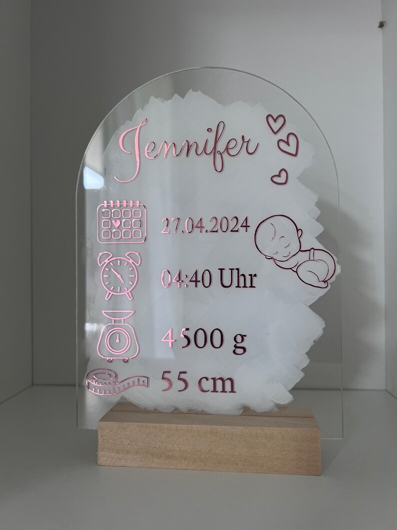 K&ouml;nnte beinhalten: Eine transparente Acryl-Plakette mit einer pinkfarbenen Inschrift, die "Jennifer" lautet und Kalender-, Uhr-, Waagen- und Ma&szlig;band-Symbole enth&auml;lt. Die Plakette enth&auml;lt auch das Datum "27.04.2024", die Uhrzeit "04:40 Uhr", das Gewicht "4500 g" und die Gr&ouml;&szlig;e "55 cm".