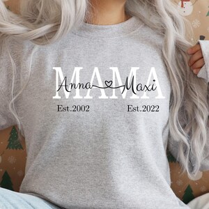 May include: Gray sweatshirt with the text "MAMA" in white with the names "Anna" and "Maxi" written in cursive on either side of the word "MAMA" with a heart symbol between the names. The text "Est. 2002" is below the name "Anna" and the text "Est. 2022" is below the name "Maxi".