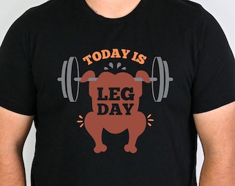 May include: Black t-shirt with a humorous graphic. The design features a turkey lifting weights, with the text "TODAY IS LEG DAY" in orange and white. The barbell is gray. A fun, fitness-themed apparel item.